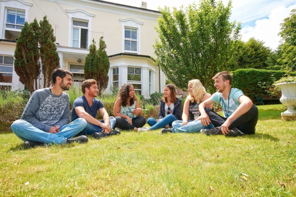 Seis estudiantes felices sentados en el césped con la escuela de inglés en Eastbourne detrás.