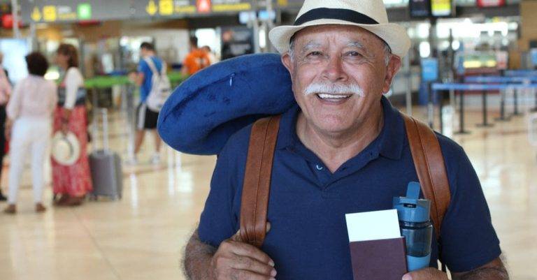 hombre mayor sonriendo en el aeropuerto con rucsac y sombrero y pasaporte en mano